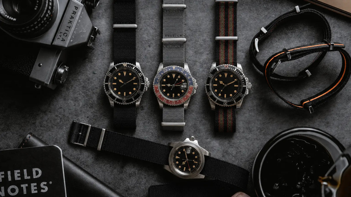Three wristwatches with black dials and mixed black and brown straps are arranged on a dark textured surface with vintage camera and notebook props.