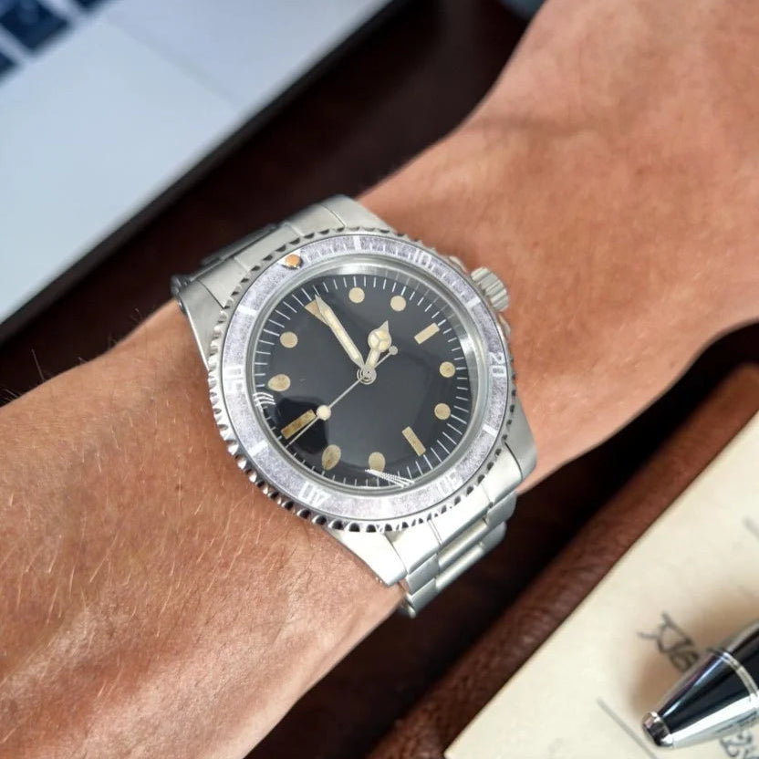 Aged ghost-bezel automatic dive watch worn on the wrist beside a notebook and laptop